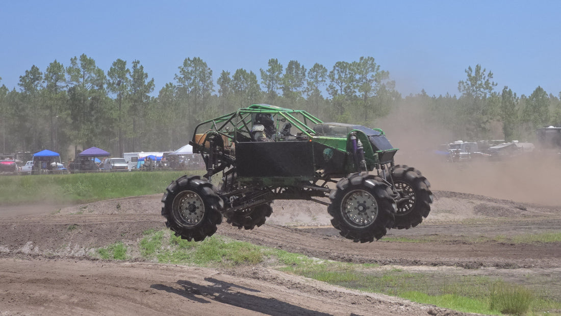 A large lime-green mega truck with massive tractor tires splashing through a mud pit under a clear blue sky at an off-road park.