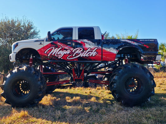 White and black monster truck named "Mega Bitch" with massive tires parked on a grassy field under a clear blue sky.