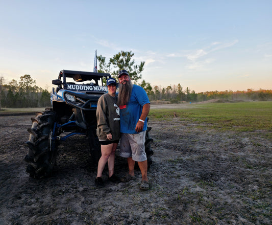 Jennifer and her husband, the founders of Mudding Murica, standing in front of a customized side-by-side vehicle in an open field at sunset.