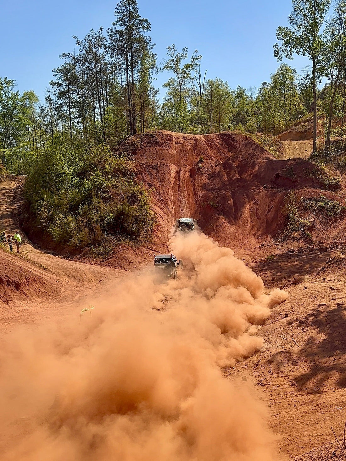“Two off-road vehicles climb a steep red dirt trail, kicking up heavy dust as they race through a rugged forested terrain