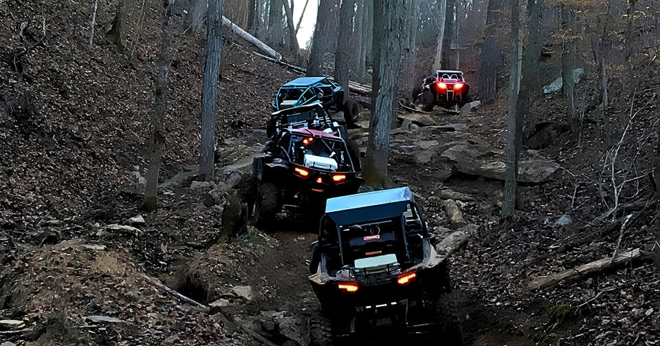 Four off-road rigs climbing a steep, rocky forest trail with bright tail lights glowing through dense trees.