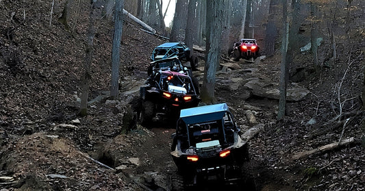 Four off-road rigs climbing a steep, rocky forest trail with bright tail lights glowing through dense trees.