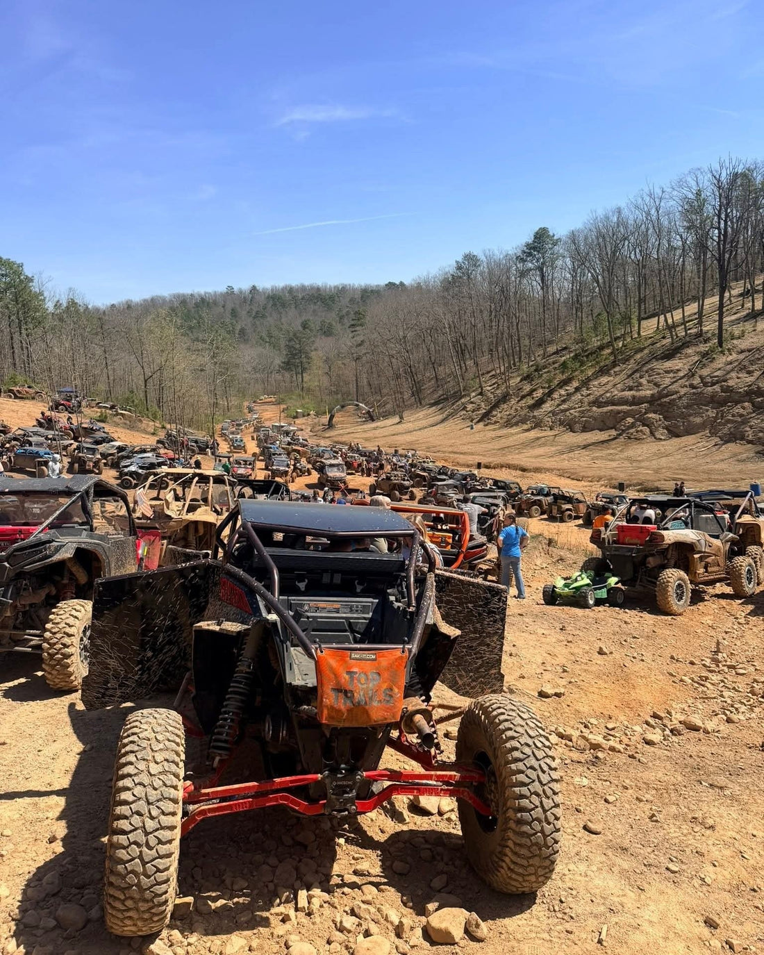 Massive line of mud‑covered rock crawlers and ATVs packed along a rugged forest trail during an off‑road event.