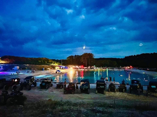 Night ride gathering with ATVs and UTVs parked by a lake, colorful lights reflecting on the water under a dark cloudy sky.