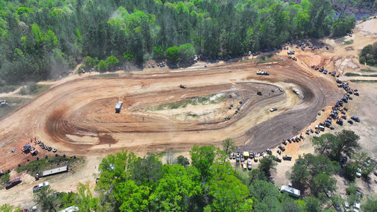 Aerial dirt racetrack with trucks racing and spectators around a muddy oval course surrounded by forested terrain.