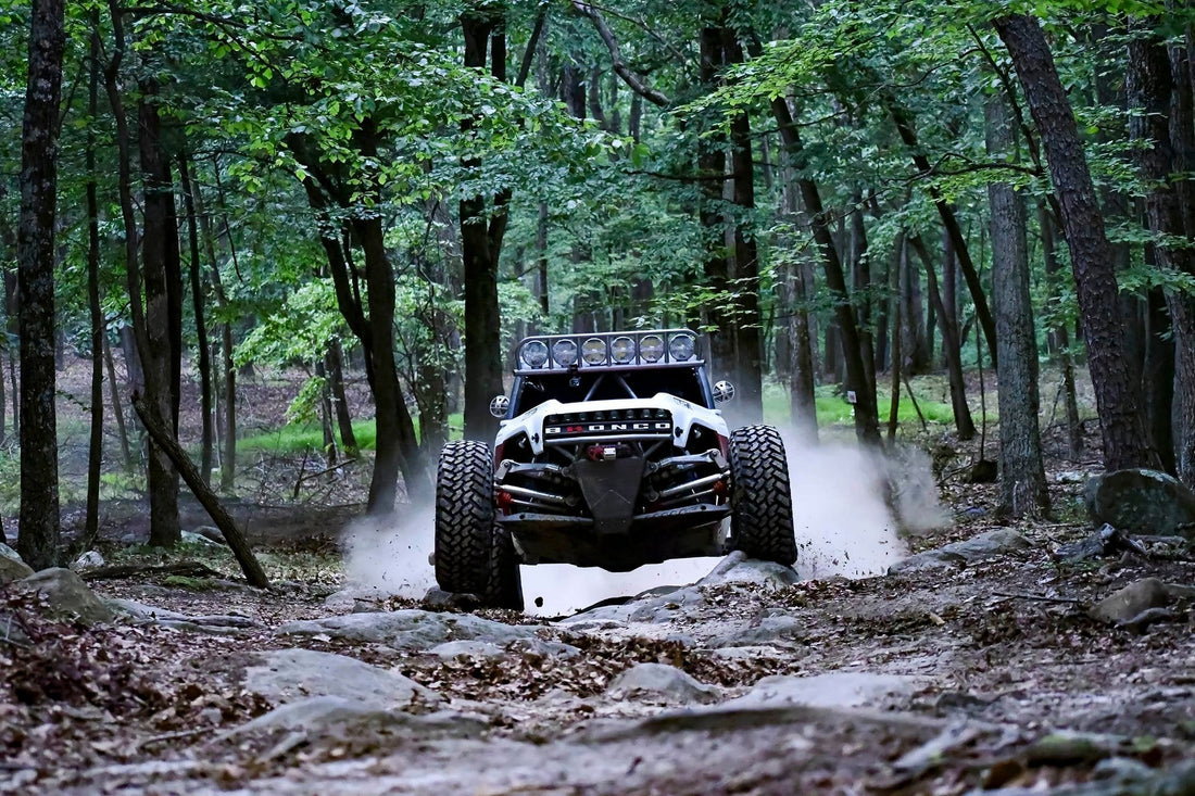 White custom Ford Bronco off-road racing truck kicking up dust while navigating a rocky trail in a dense forest.