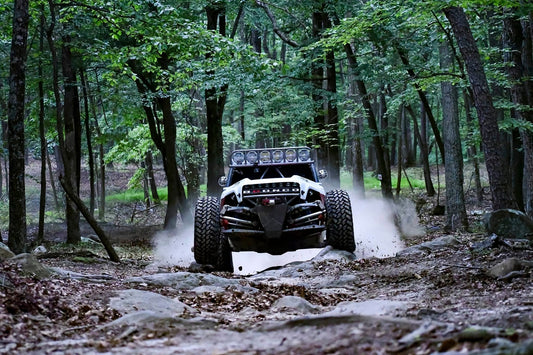 White custom Ford Bronco off-road racing truck kicking up dust while navigating a rocky trail in a dense forest.