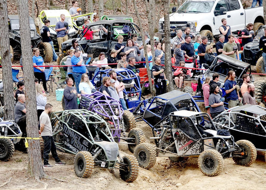Crowd gathered around rock buggies and off‑road rigs on a wooded dirt trail during an outdoor event with spectators and drivers.