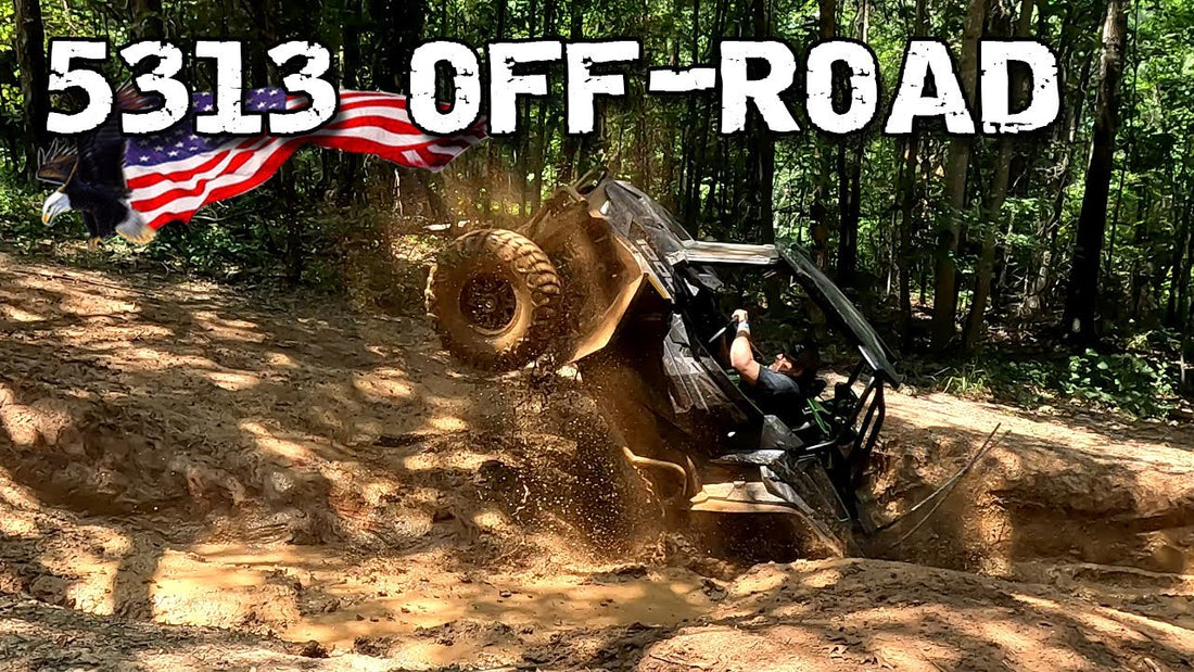 "A side-by-side UTV performing a steep, muddy wheelie in a wooded trench with '5313 OFF-ROAD' and American flag graphics."