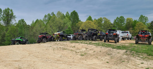 Line of UTVs parked on a dirt hill with trucks and trailers behind them at a forested off‑road staging area
