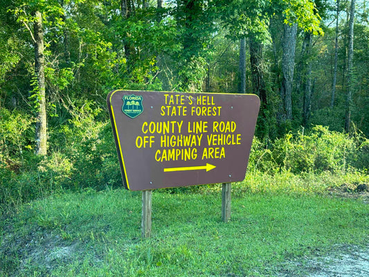 "Roadside sign for Tate's Hell State Forest County Line Road OHV camping area surrounded by lush Florida pine trees."